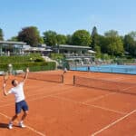 Joueurs de tennis sur terre battue devant un club-house moderne et verdoyant à Boulogne-Billancourt.