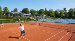 Joueurs de tennis sur terre battue devant un club-house moderne et verdoyant à Boulogne-Billancourt.