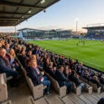 Vue latérale des tribunes d'un stade de football remplies de supporters souriants lors d'un match en soirée.