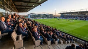 Vue latérale des tribunes d'un stade de football remplies de supporters souriants lors d'un match en soirée.