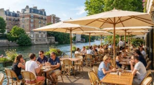 Clients attablés en terrasse au bord de l'eau, sous des parasols beige, avec des immeubles parisiens en arrière-plan.