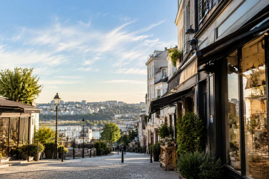 Rue commerçante pavée avec boutiques sous un ciel ensoleillé, vue en plongée sur une ville en arrière-plan.