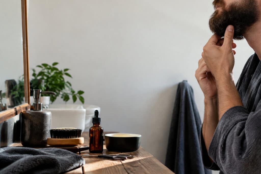 Un homme en peignoir examine sa barbe devant un miroir avec des produits d'entretien sur une table en bois.
