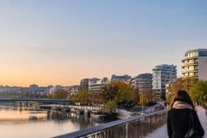 Vue de dos d'une personne marchant sur un pont face à la Seine et aux immeubles modernes de Boulogne-Billancourt au crépuscule.