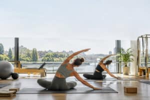 Deux femmes pratiquent des étirements Pilates sur des tapis dans un studio lumineux avec vue panoramique sur une rivière.