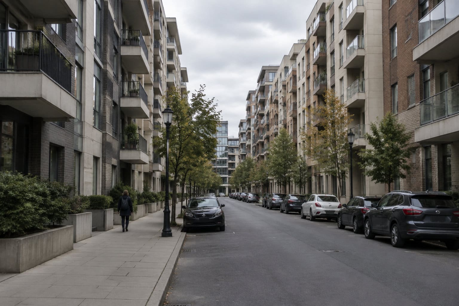 Une rue bordée d’immeubles résidentiels, d’arbres et de voitures garées, avec une personne qui marche sur le trottoir sous un ciel nuageux.