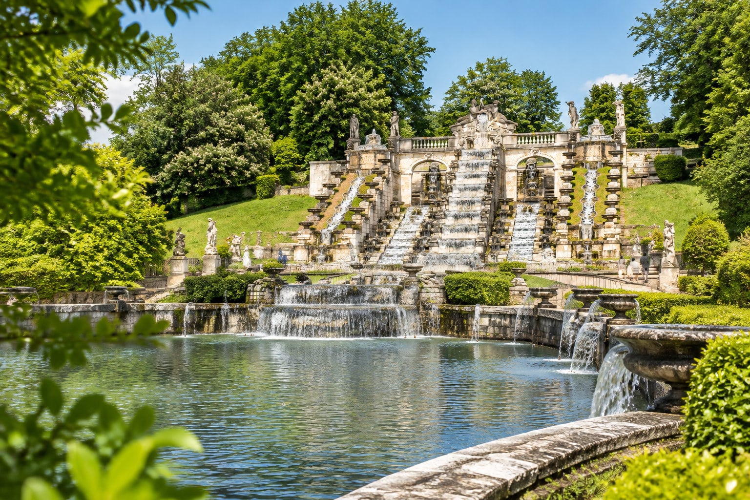 Une grande cascade ornementale en pierre dévale plusieurs bassins dans un jardin verdoyant, entourée de statues et d’arbres.